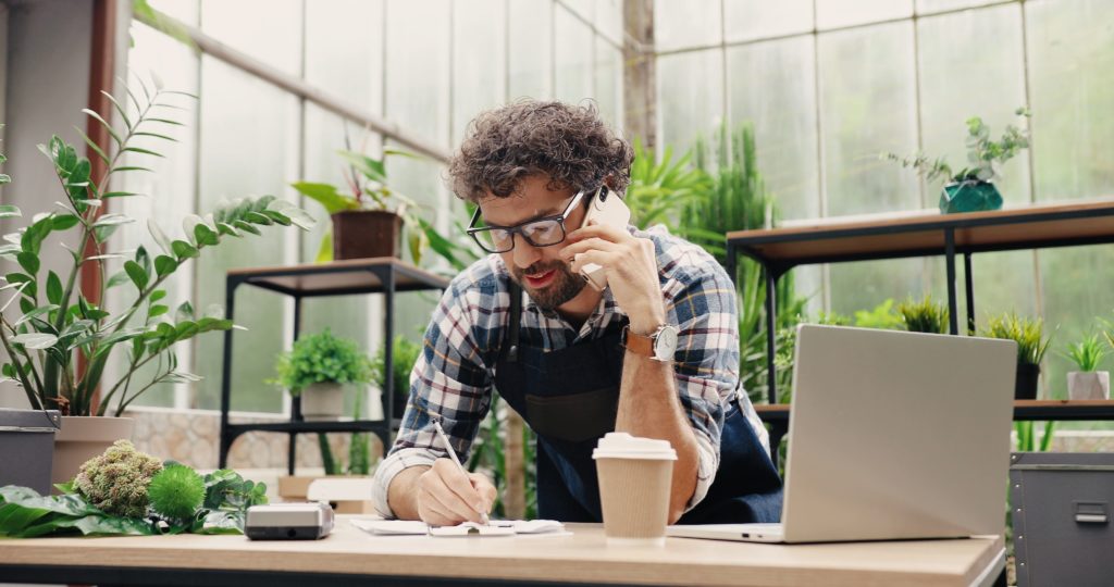 Happy businessman talking on cellphone while standing in apron in small floral center and writing down order details. Joyful male florist calling on smartphone at work.